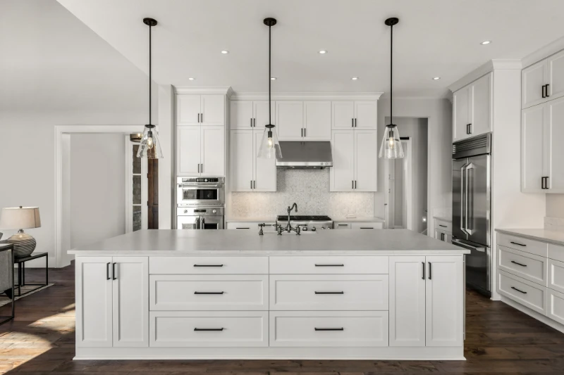 Modern white kitchen with large island featuring drawers, pendant lighting, stainless steel appliances, white cabinets, and dark hardware, set on a wooden floor with a clean, minimalist design.