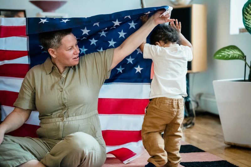 A woman and child playfully interact under an American flag in a cozy indoor setting.