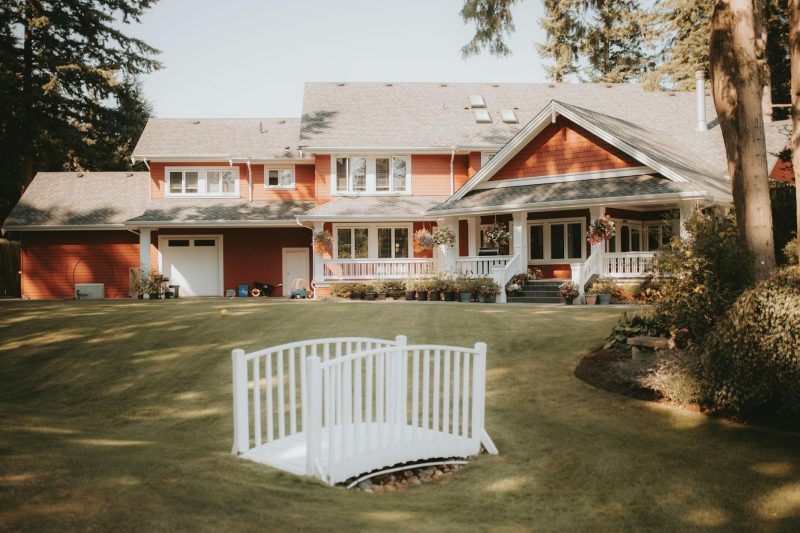 Charming red house with a large yard and a white bridge, set against a serene landscape.