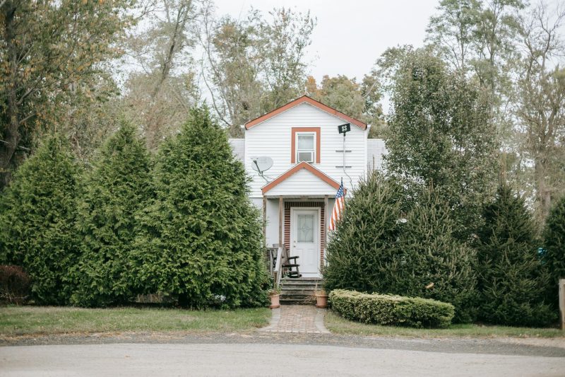 Cottage facade with an American flag, surrounded by fir trees near an asphalt road in a suburban setting.