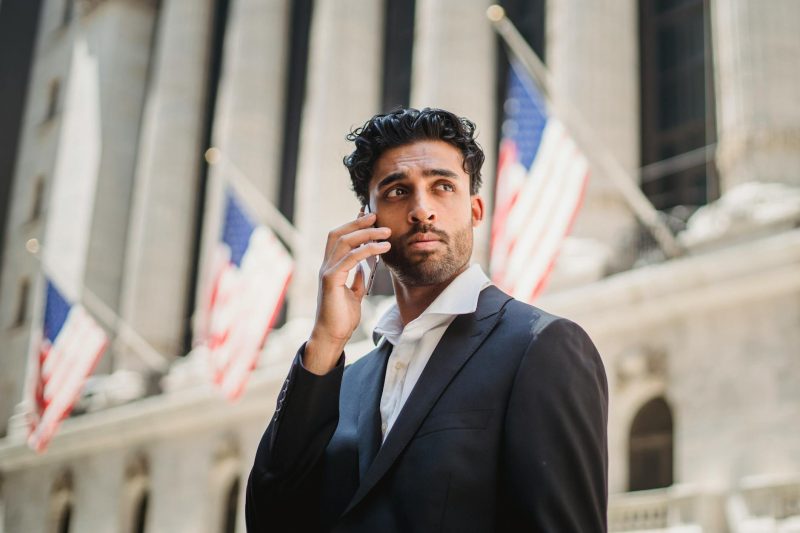 Businessman in a black suit talking on a mobile phone outside a historic building with flags.