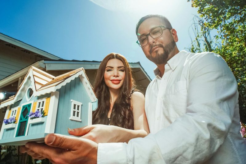 A happy couple outdoors holds a miniature house model, smiling and celebrating home ownership.
