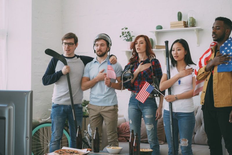 Diverse friends cheer while watching sports on TV, surrounded by American flags in a cozy indoor setting.