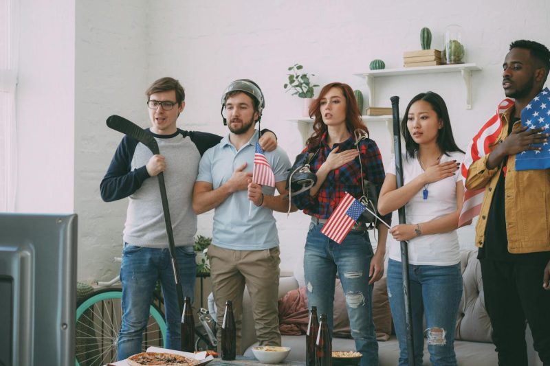Diverse friends gathered indoors, watching sports on TV, with American flags decorating the walls.