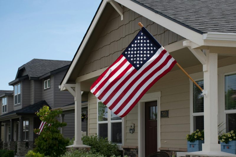 A vibrant American flag flutters outside a stylish suburban house surrounded by greenery in daylight.