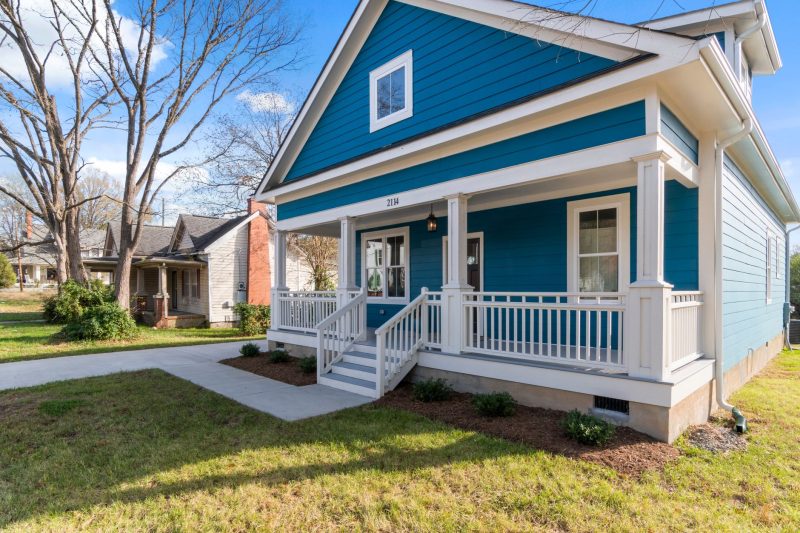 Cozy blue suburban house with a white porch, surrounded by lush greenery on a clear day.