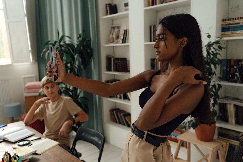 A woman takes a selfie in a stylish, plant-filled study room with a young boy observing her.