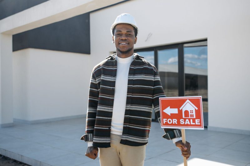 Young black male realtor smiles while holding a for sale sign in front of a modern house.