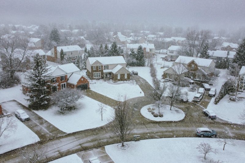 Aerial view of a snow-covered suburban neighborhood with rooftops, streets, and scattered trees.